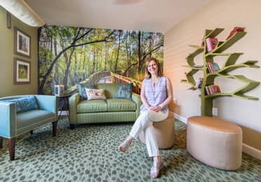 Woman sitting in a room with forrest wallpaper and books on a shelf.